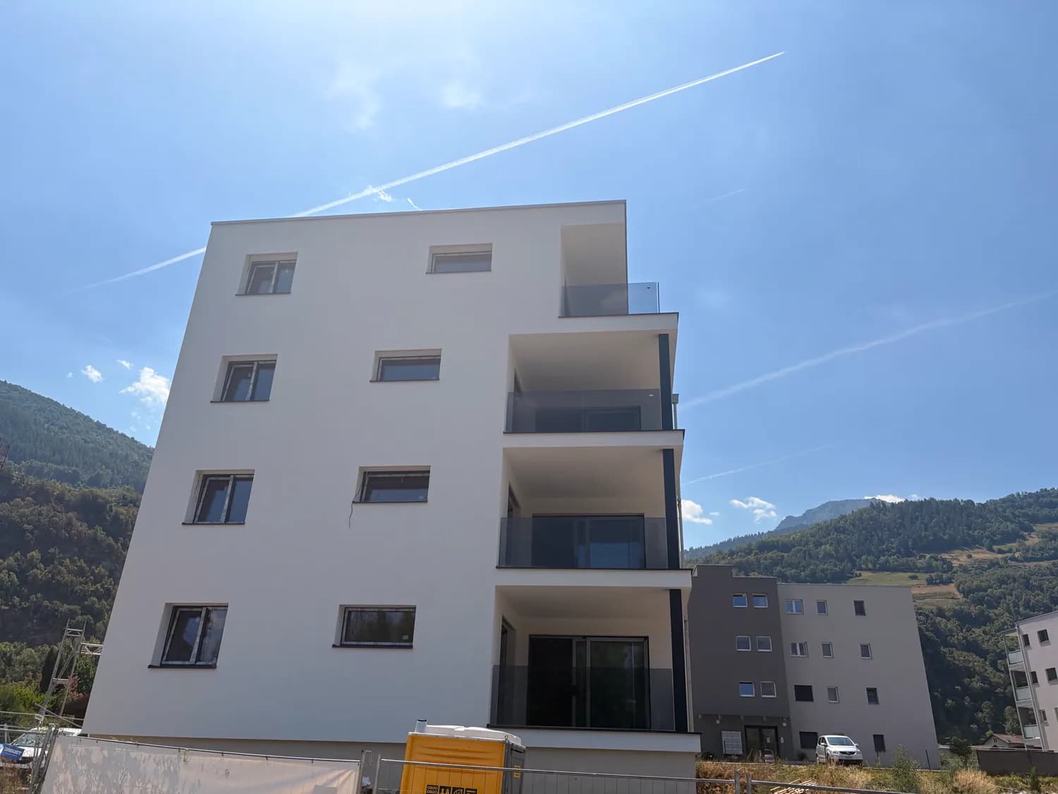 Neubau mit Balkonen und Geländern aus Rauchglas, bewaldeter Hang im Hintergrund
